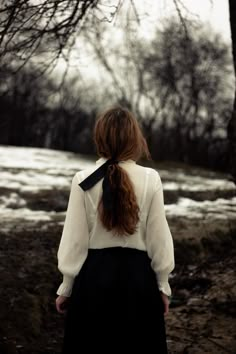 a woman with long red hair wearing a white shirt and black skirt is standing in the woods