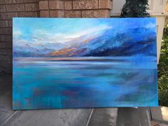 an acrylic painting of mountains and blue water on a wooden easel in front of a brick wall