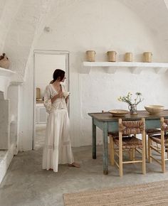 a woman standing in a room next to a table with chairs and vases on it