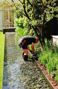 a man kneeling down to pick up rocks in the water from a small stream that runs between two buildings