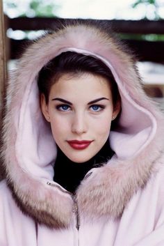 a woman with blue eyes wearing a pink fur coat and hood over her head is looking at the camera