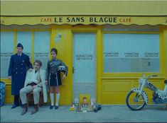 three people sitting on a bench in front of a yellow building with french words written on it