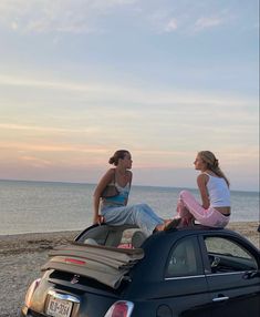 two women sitting on top of a car near the ocean