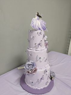 a three tiered wedding cake with purple flowers and butterflies on the top, sitting on a white table cloth