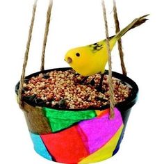 a yellow bird sitting on top of a colorful pot filled with birdseed and hanging from strings