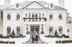 two people standing in front of a large white house with wreaths on the windows