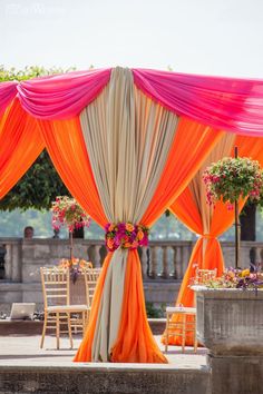 an outdoor wedding setup with orange and pink draping on the top, flowers in vases at the bottom