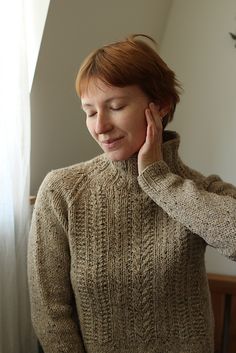 the woman is wearing a sweater and holding her hand to her ear as she stands in front of a window