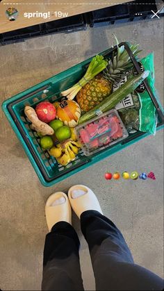 a person standing next to a plastic container filled with fruits and vegetables on the floor