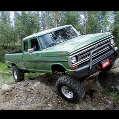 a green pick up truck driving on top of a dirt hill in the woods with trees behind it