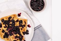 waffles with blueberries are on a white plate next to a bowl of berries