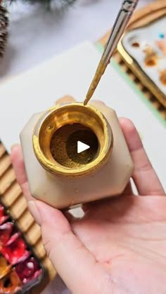 a person holding a small gold cup with a spoon in it and some other items on the table