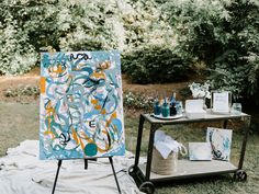 an easel sitting on top of a grass covered field next to a table filled with art supplies