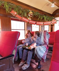 two women in pajamas are sitting on a train car with christmas decorations hanging from the ceiling