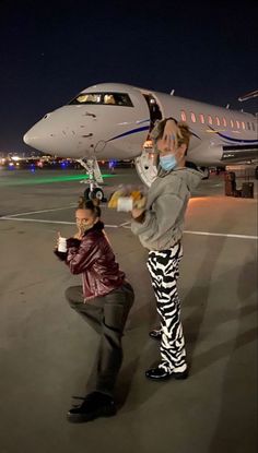 two people standing in front of an airplane on the tarmac at night, one holding up a camera