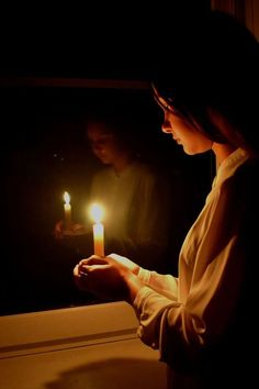 a woman holding a lit candle in her hand and looking out the window at another person