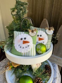 two snowmen sitting on top of a three tiered tray with green ornaments and pine cones