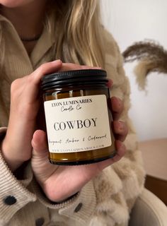 a woman holding a jar of cowboy soy candle in front of her face while wearing a scarf