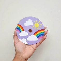 a hand holding up a purple paper plate with rainbows and clouds painted on it