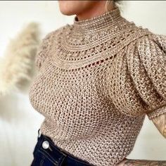 a woman with tattoos on her arm wearing a knitted sweater and denim jeans, standing in front of a white wall