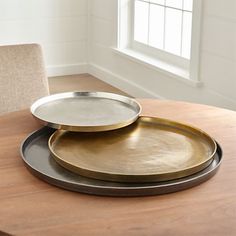 two gold and silver plates sitting on top of a wooden table next to a window