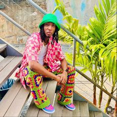 a man sitting on top of a wooden bench wearing colorful pants and a green hat