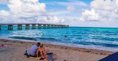 two people are sitting on the beach by the water and under an overpass with a pier in the background