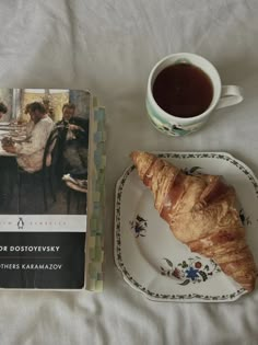 a croissant on a plate next to a coffee cup and book about food