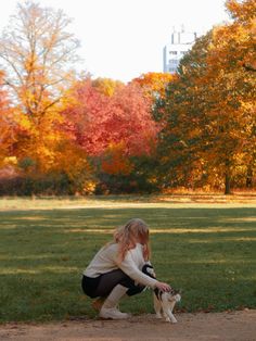 a woman kneeling down petting a dog in a field with trees and buildings in the background