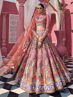 a woman standing in front of a pink wall wearing a colorful lehenga set