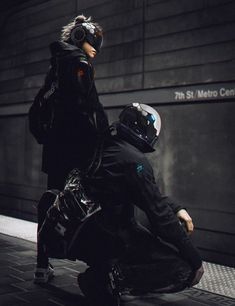 two people in black jackets and helmets sitting on the ground next to a wall with a sign