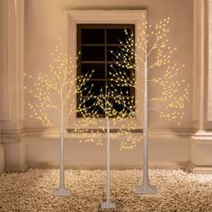 two white trees with yellow lights in front of a window on carpeted floor next to windowsill