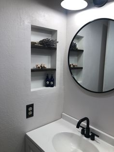 a bathroom sink under a round mirror next to a shelf with bottles and soaps on it