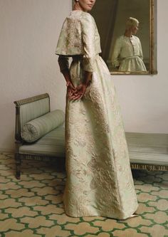 a woman standing in front of a mirror wearing a white dress with gold embroidery on it