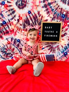 a baby sitting on a red blanket holding a sign that says, baby you're a firework