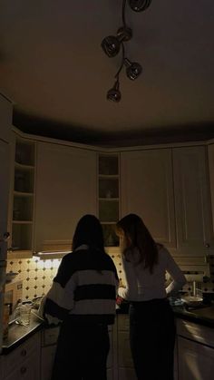 two people standing in a kitchen looking at something on the counter top and ceiling lights above them