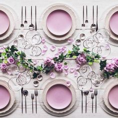 the table is set with pink plates, silverware and purple flowers on top of it