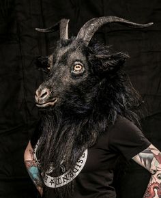 a man wearing a goat mask with tattoos on his arm and chest, standing in front of a black background