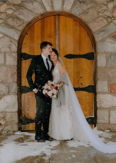 a bride and groom standing in front of an arched door with snow falling on them