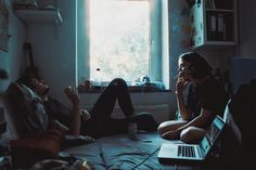 two people sitting on the floor in front of a window with their laptops open