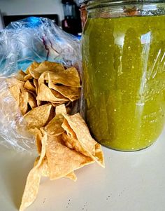 a glass jar filled with green liquid next to some tortilla chips on a table