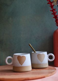 two coffee mugs with hearts painted on them sitting on a wooden tray next to a red vase