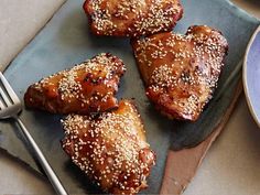 four pieces of chicken with sesame seeds on a plate next to a fork and bowl