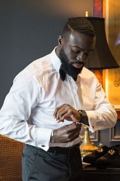 a man in a white shirt and bow tie looking at his watch while standing next to some shoes