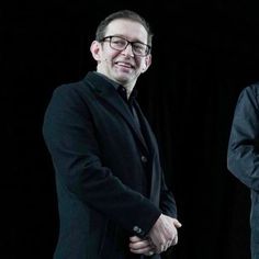 two men standing next to each other in front of a black background with one man wearing glasses