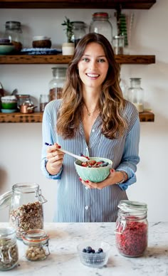 a woman holding a bowl of food in front of some jars and spoons on a table