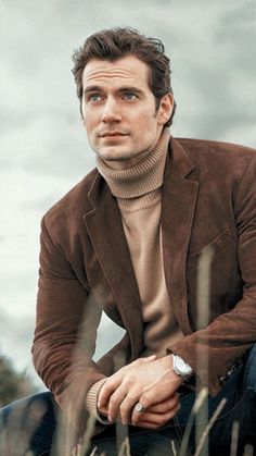 a man sitting in the grass wearing a brown jacket and turtle neck sweater with his hands on his knees