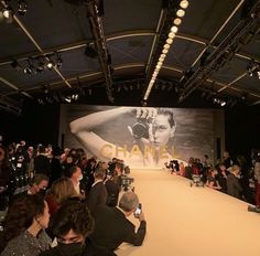 a crowd of people sitting in front of a chanel advertisement on the wall at a fashion show