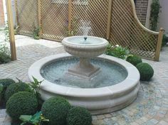 a water fountain in the middle of a brick patio surrounded by greenery and shrubbery