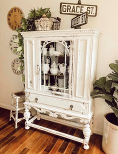 a white china cabinet sitting on top of a wooden floor next to a potted plant